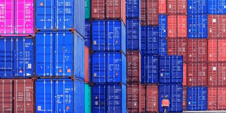 Stacked shipping containers. Most of them are blue, a few are crimson and in the top left corner is a pink shipping container. A man on a bicycle is in the foreground.