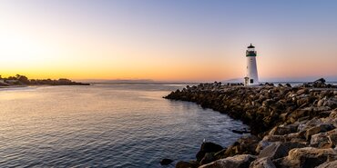 A white lighthouse sits atop a rocky sea wall as the sun sets behind it. The sky is a light blue with streaks of pink and yellow