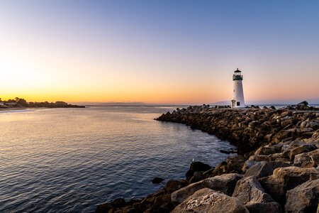 A white lighthouse sits atop a rocky sea wall as the sun sets behind it. The sky is a light blue with streaks of pink and yellow