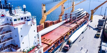 a yellow crane lifting a huge wind turbine blade onto a white a red ship in a blue sea.