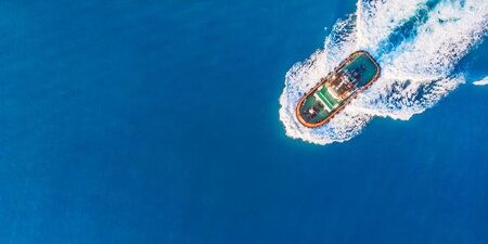 a tug boat from above sails across deep blue water with a white wake behind