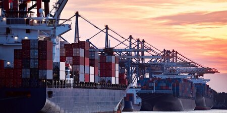 Three large stacked container ships in a row at dusk. Port cranes and some of the sea is also visible