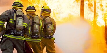 Three firefighters tackle a blaze.The image is taken behind the three firefighters who are spraying the fire with a hose.